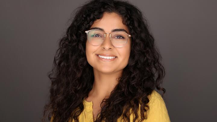 Woman with clear frame glasses, brown curly hair, and a yellow top.