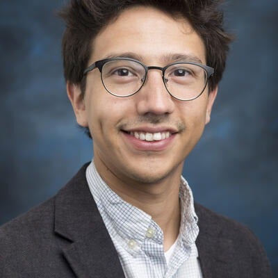 Javier Garibay is of lighter complexion with short brown hair, wearing glasses, and he is wearing a dark grey blazer against a blue backdrop.